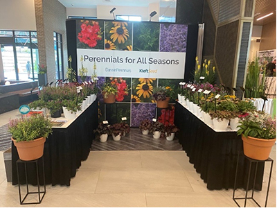 Two tables of perennials flank a backwall of Perennials for All Seasons signage.